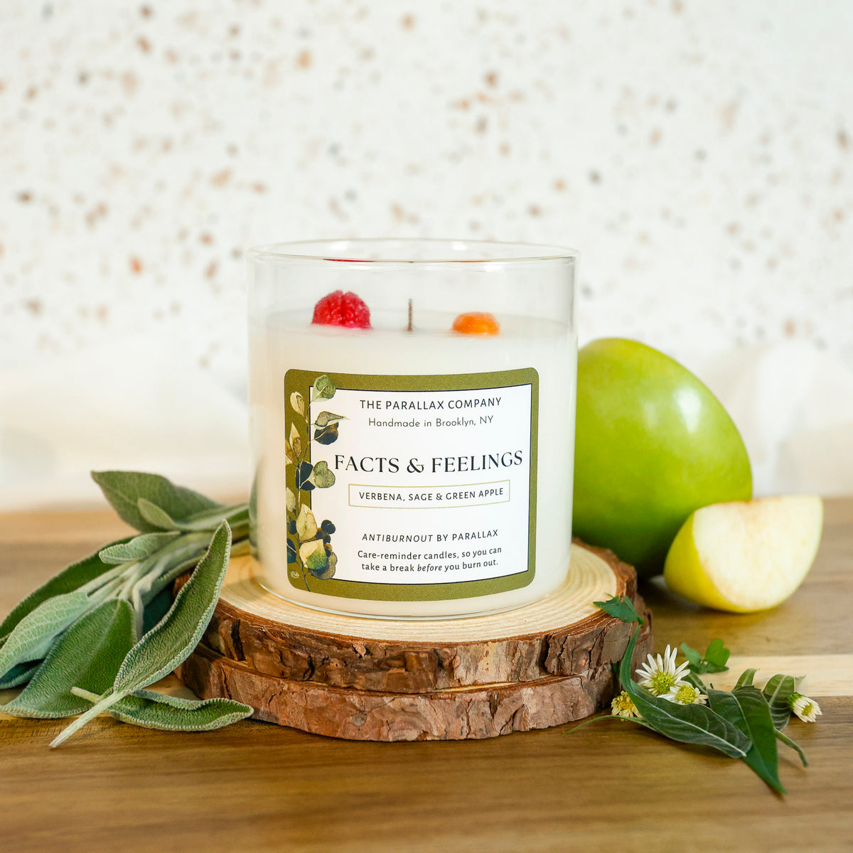 A glass jar candle with white wax sits on a round wood slice, surrounded by sage leaves, a green apple, and a sliced apple on a wooden surface. The candle label reads 'Facts & Feelings' by The Parallax Company.
