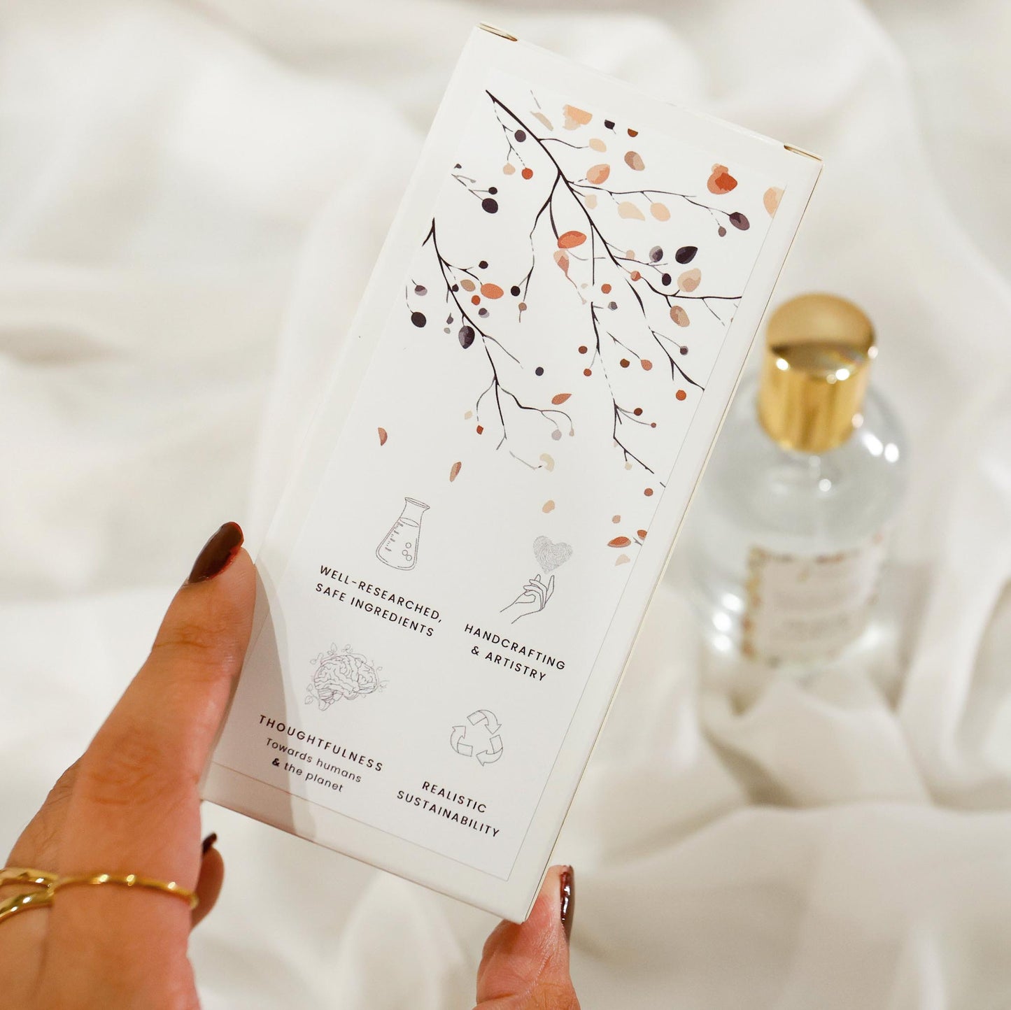 Hand holding a room spray product packaging box with decorative pattern design on a soft white background and brand value icons: Well-researched ingredients, Handcrafting & Artistry, Thoughtfulness towards humans & the planet, Realistic Sustainability