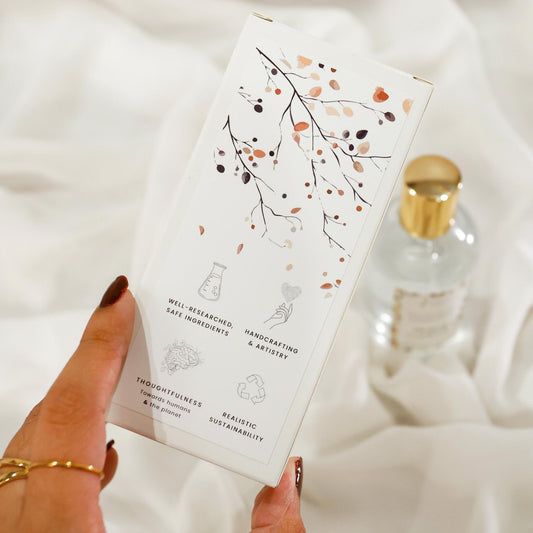 Hand holding a room spray product packaging box with decorative pattern design on a soft white background and brand value icons: Well-researched ingredients, Handcrafting & Artistry, Thoughtfulness towards humans & the planet, Realistic Sustainability