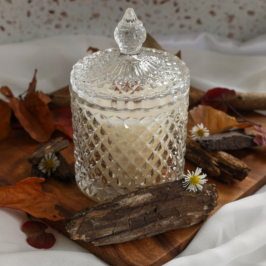 A clear cut-glass candle jar with a matching lid, filled with a white wax and wooden wick, styled with dried Fall leaves, wood bark, and chamomile flowers.