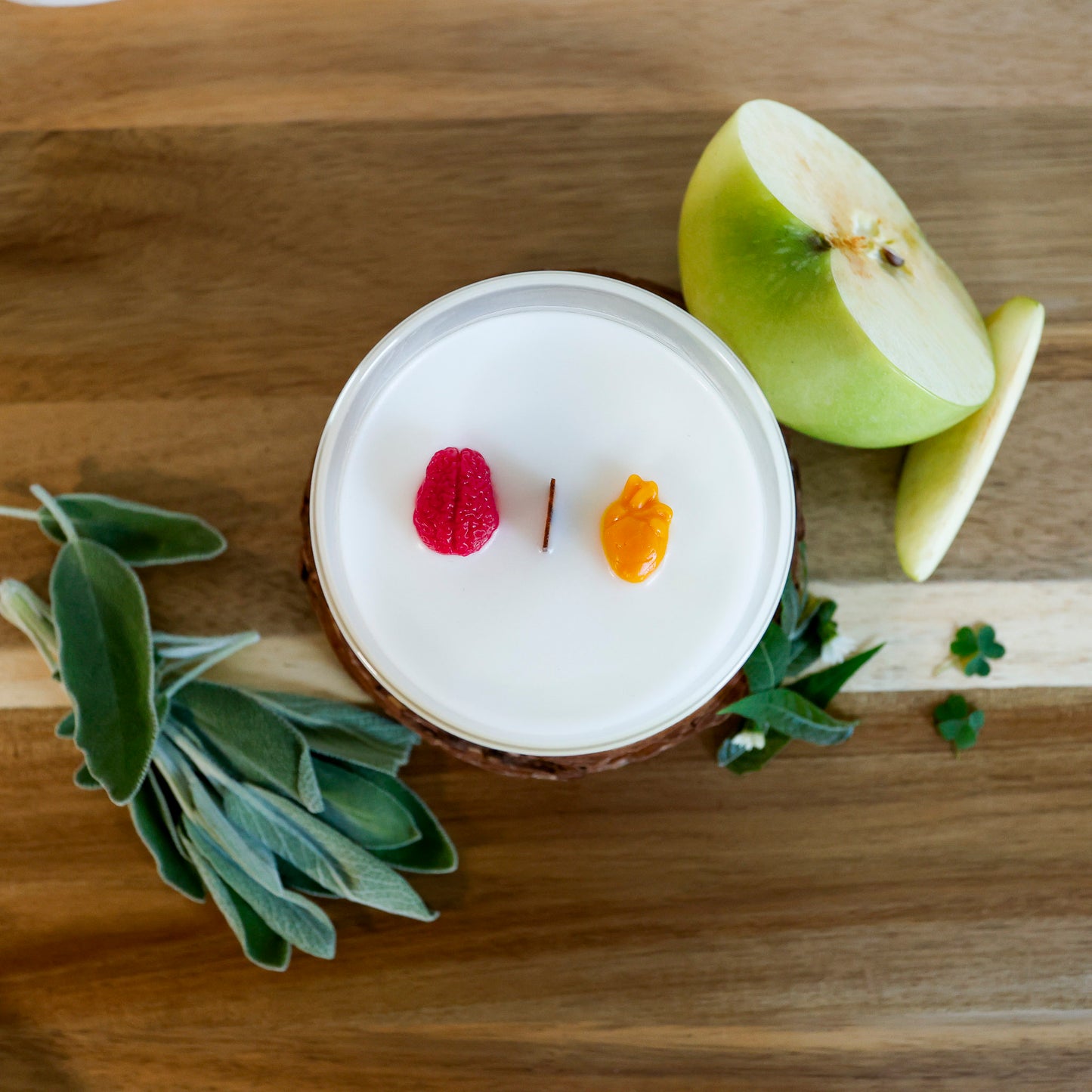 A round wooden wick candle in a clear glass jar, decorated with a 3D miniature human brain and a small orange human heart, sits amidst a sliced green apple, sage leaves, and lemon verbena.