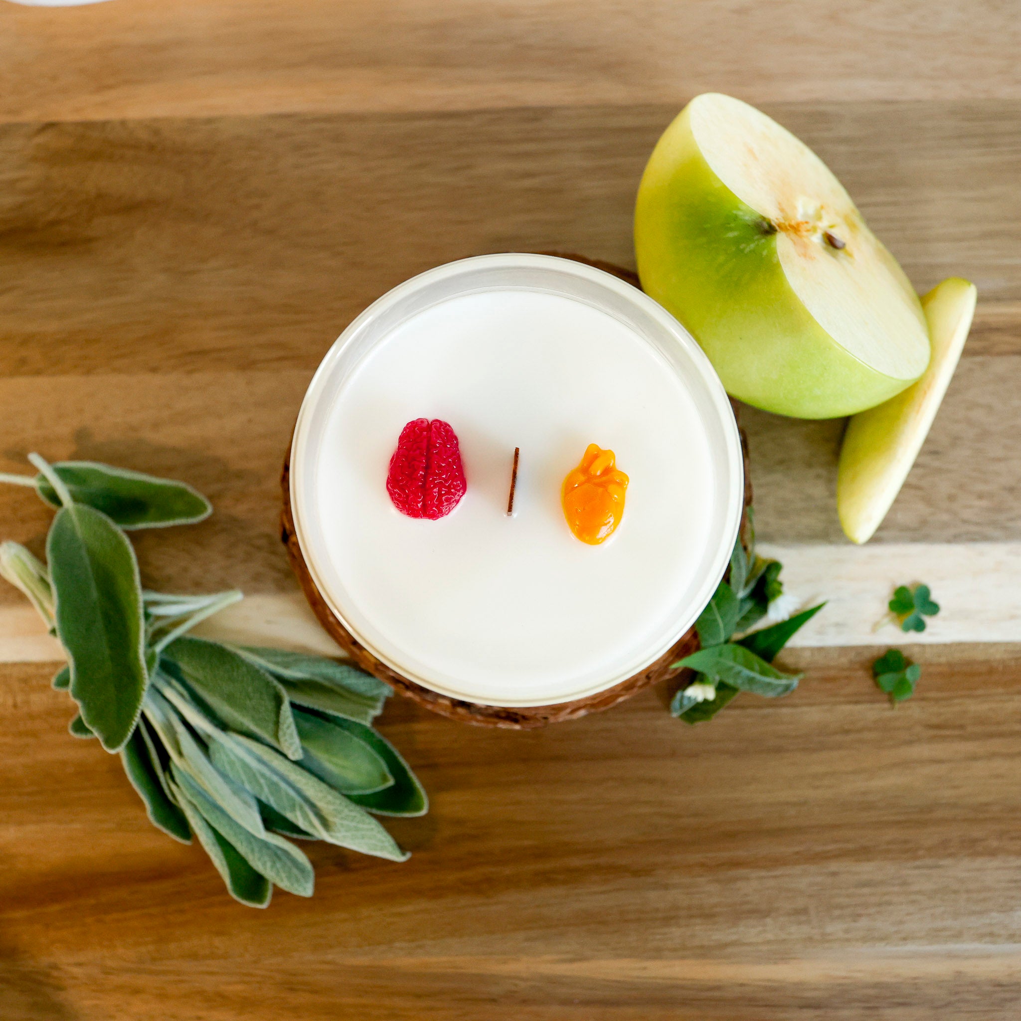 A round wooden wick candle in a clear glass jar, decorated with a 3D miniature human brain and a small orange human heart, sits amidst a sliced green apple, sage leaves, and lemon verbena.