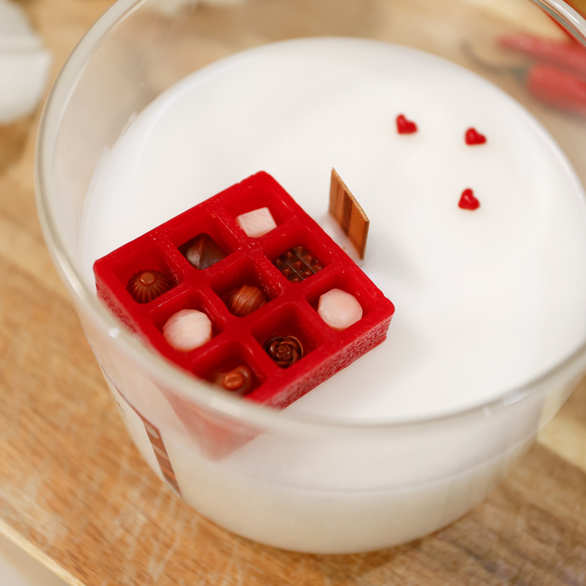 Red box with assorted chocolates in wax with small hearts on scented wood wick candle, surrounded by roses and chillies.