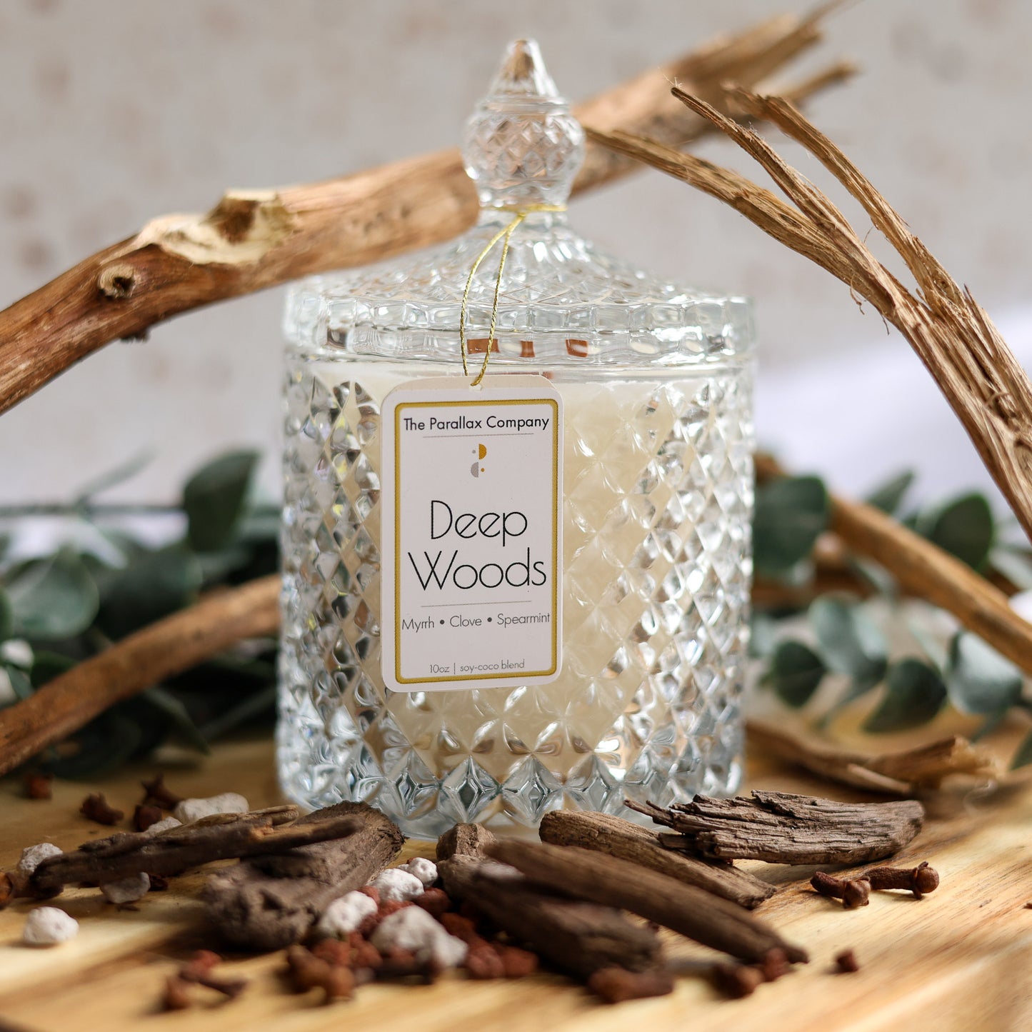 Decorative glass candle jar with a 'Deep Woods' white and gold scent label, placed with woody bark, tree branches and eucalyptus leaves.