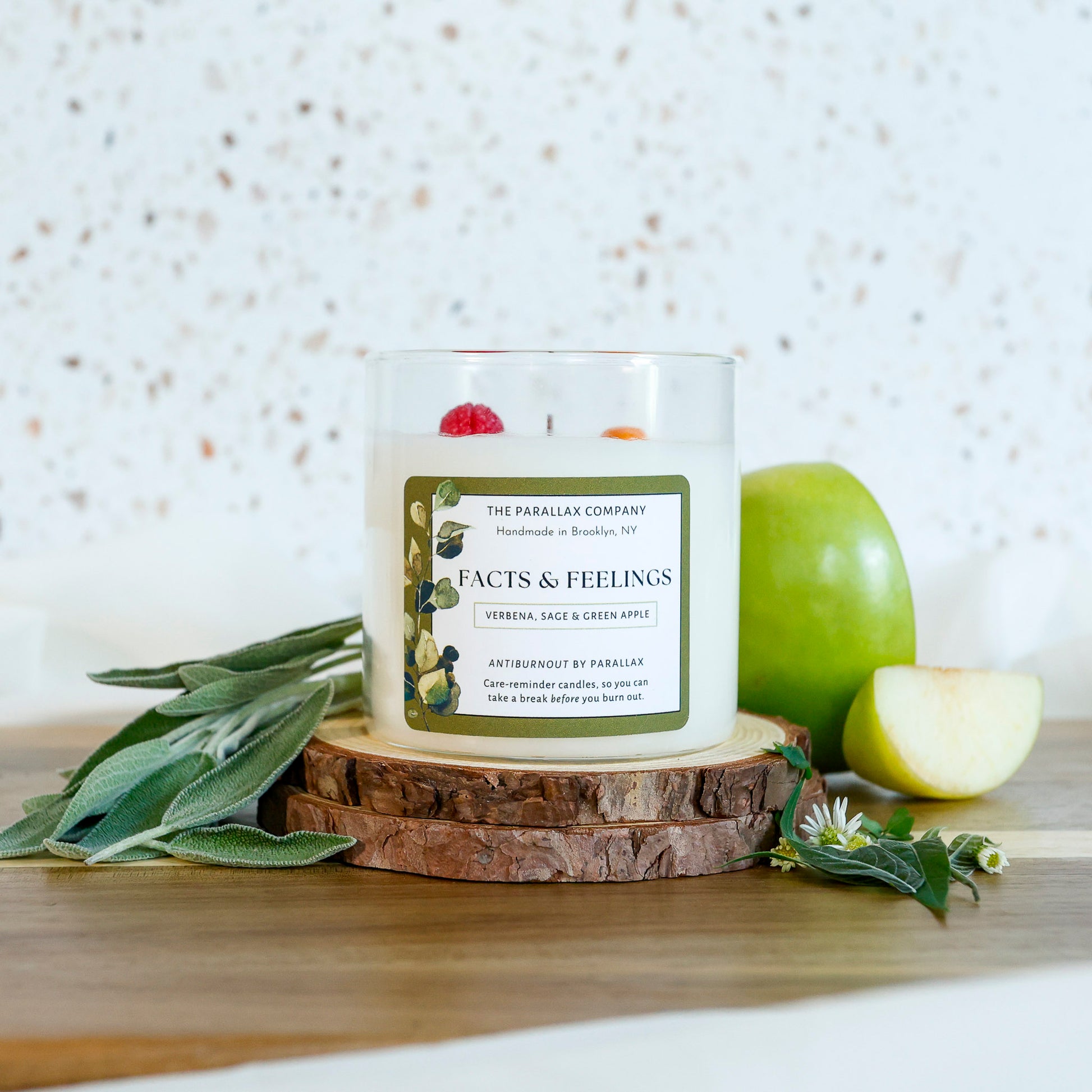 A glass jar candle labeled 'Facts & Feelings' by The Parallax Company sits on  rustic wood bark The candle is styled with sage leaves, a green apple, and lemon verbena on a wooden board against a terrazzo wall.