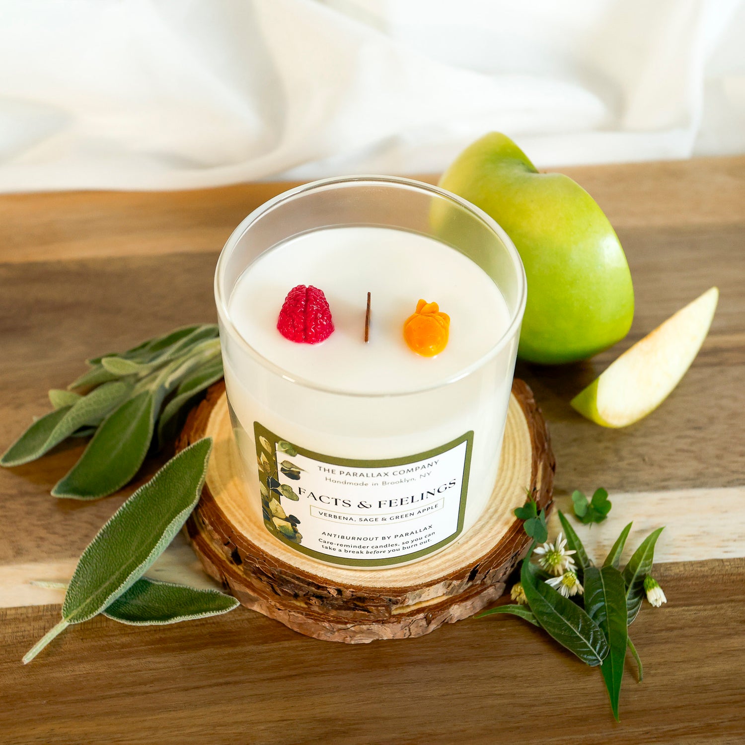 An anatomical heart and brain sculpture sit on white soy wax candle in a clear glass jar, on a wooden table, styled with sage leaves, a green apple, and a slice of apple. The label reads 'The Parallax Company' and 'Facts & Feelings'.