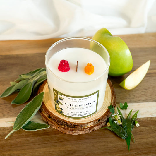 An anatomical heart and brain sculpture sit on white soy wax candle in a clear glass jar, on a wooden table, styled with sage leaves, a green apple, and a slice of apple. The label reads 'The Parallax Company' and 'Facts & Feelings'.