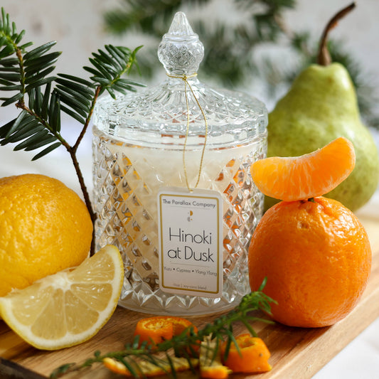 A crystal-cut glass candle jar with a label reading 'Hinoki at Dusk', surrounded by citrus fruits and woody elements, with Japanese cypress in the background.