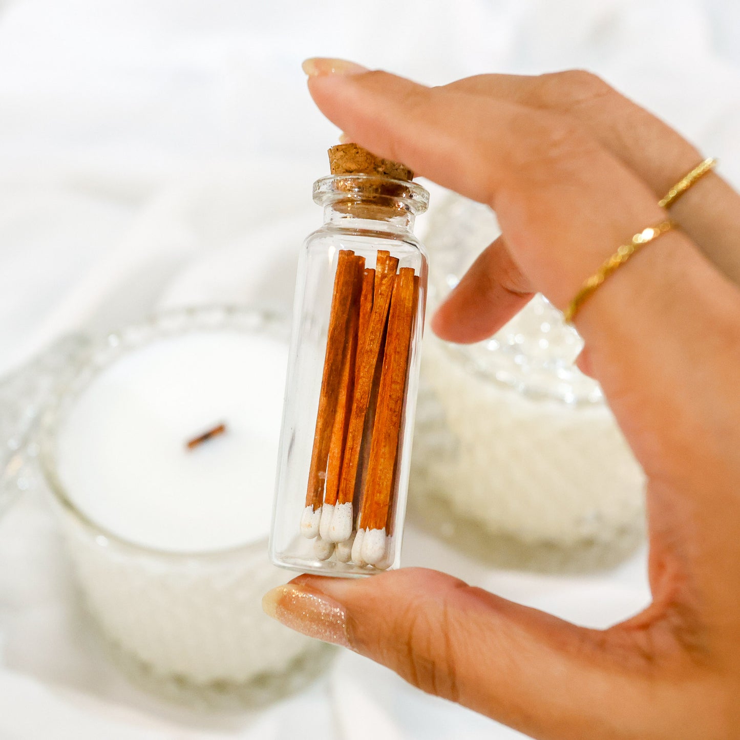 Matchsticks in a a small glass bottle vial with a cork stopper in front of a wooden wick candle.