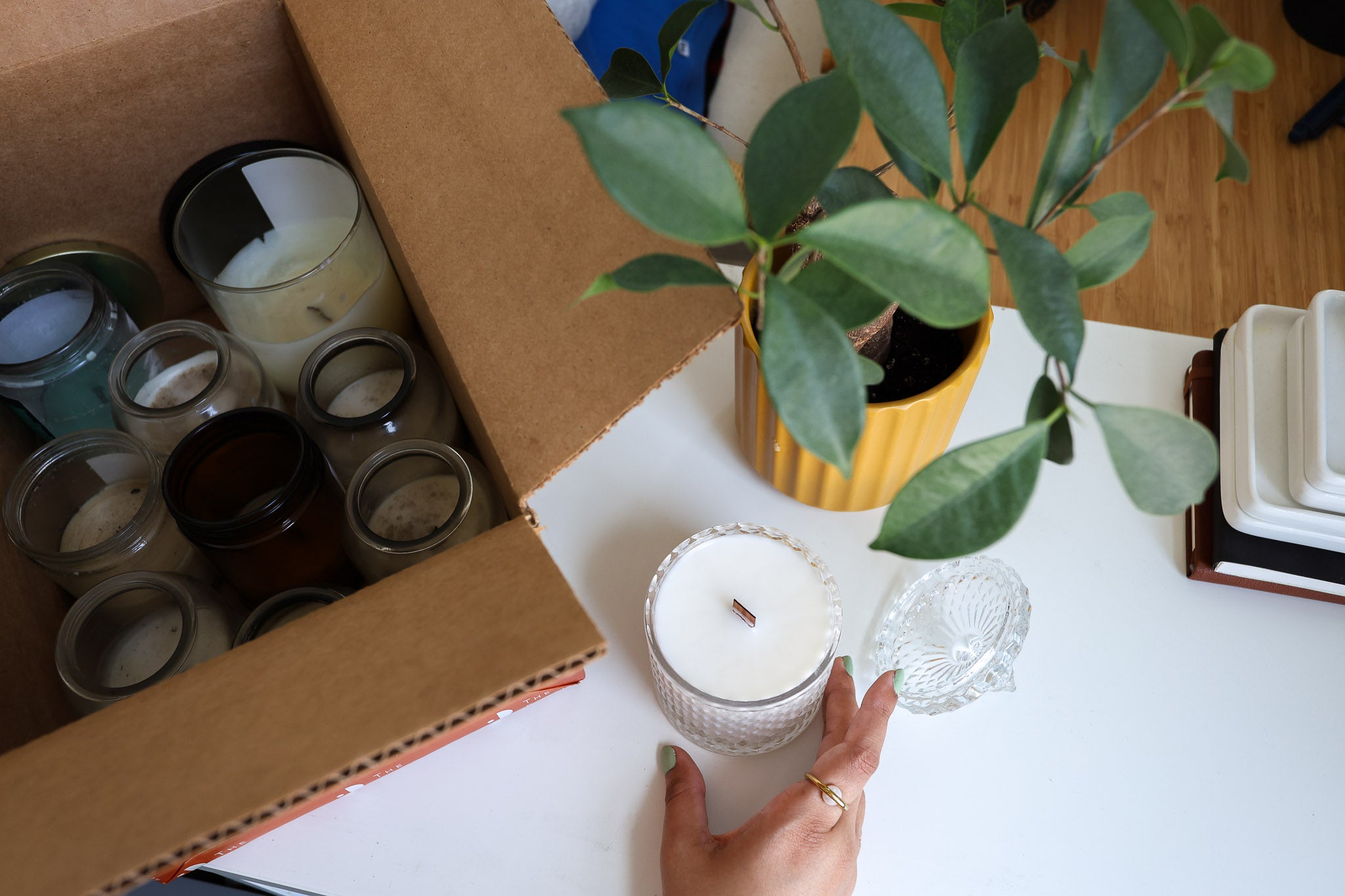 Hand holding a candle next to an open box of used candles empties on a table with a plant in the background.