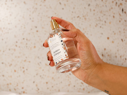 Hand with maroon fall autumn nails holding a luxury clear glass and gold bottle of room and linen spray mist against a terrazzo background