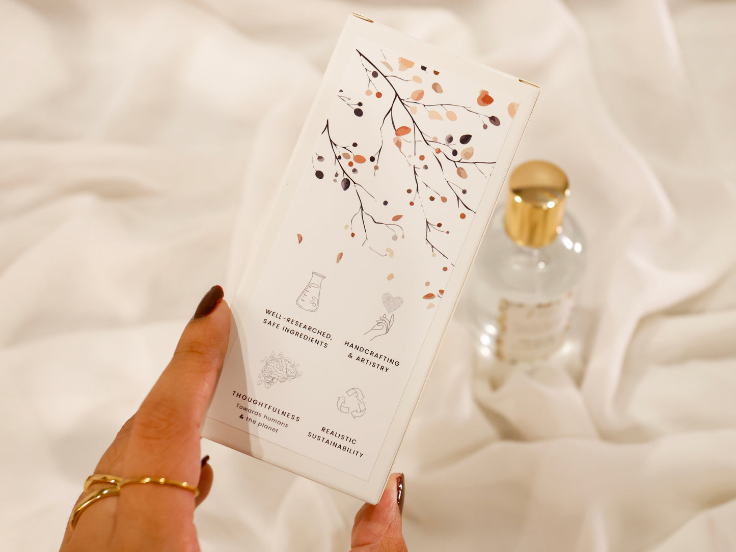 Hand holding a room spray product packaging box with decorative pattern design on a soft white background and brand value icons: Well-researched ingredients, Handcrafting & Artistry, Thoughtfulness towards humans & the planet, Realistic Sustainability
