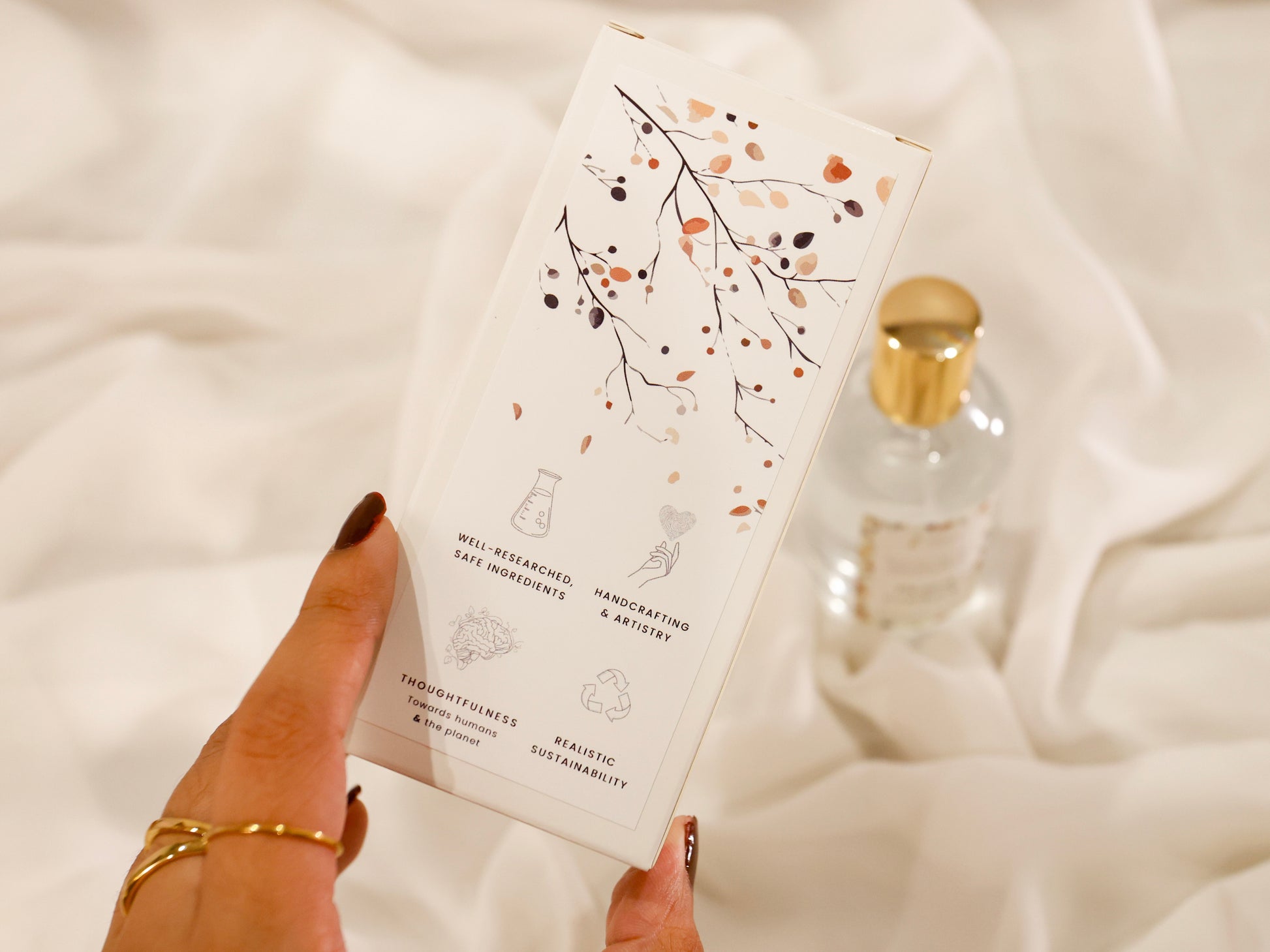 Hand holding a room spray product packaging box with decorative pattern design on a soft white background and brand value icons: Well-researched ingredients, Handcrafting & Artistry, Thoughtfulness towards humans & the planet, Realistic Sustainability