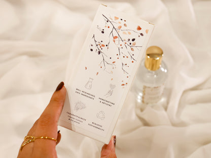 Hand holding a room spray product packaging box with decorative pattern design on a soft white background and brand value icons: Well-researched ingredients, Handcrafting & Artistry, Thoughtfulness towards humans & the planet, Realistic Sustainability