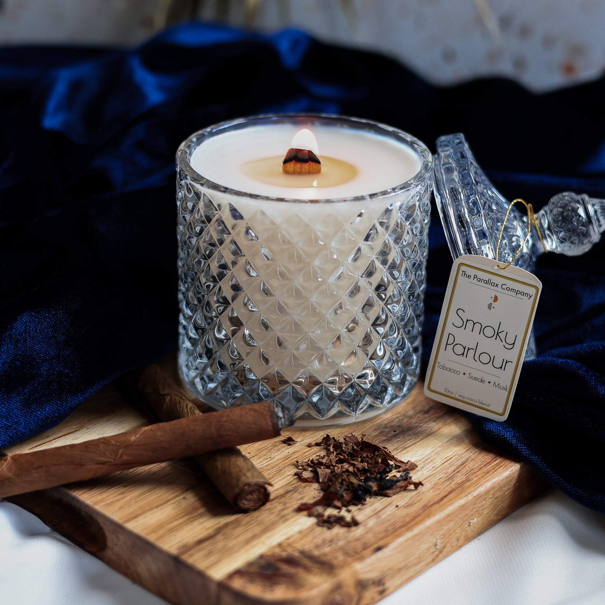 Wooden wick candle in a crystal textured glass jar, with cigars and tobacco leaves on a wooden board, against a blue luxurious velvet fabric background.