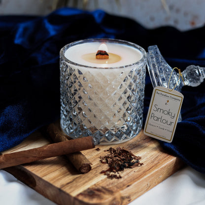 Wooden wick candle in a crystal textured glass jar, with cigars and tobacco leaves on a wooden board, against a blue luxurious velvet fabric background.