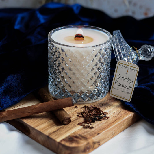 Wooden-wick man scent candle in a decorative glass jar on a wooden board with cigars, suede, and tobacco leaves, named 'Smoky Parlour'.