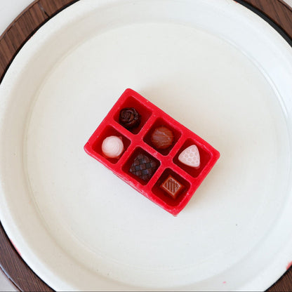 Mini wax melt shaped like a red box of assorted chocolates, on a white candlewarmer dish.