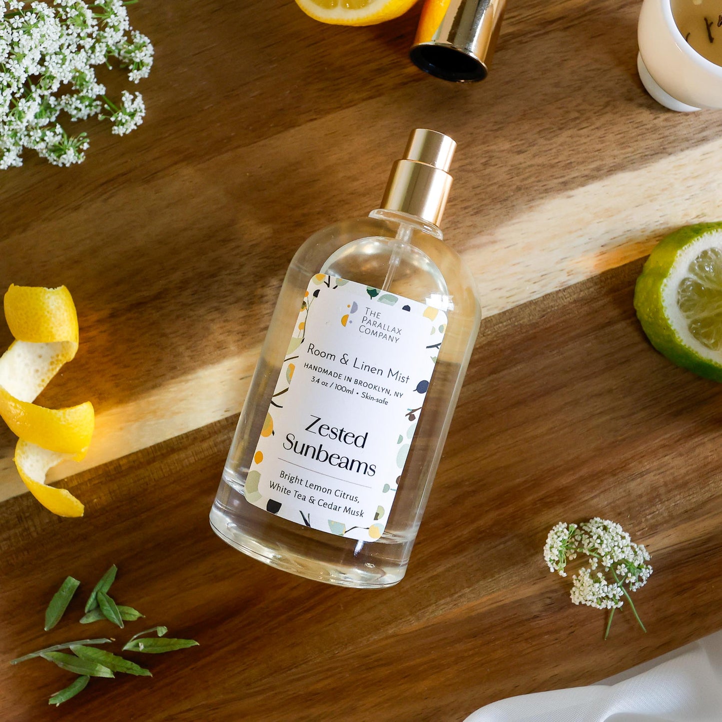 Room and linen mist bottle labeled 'Zested Sunbeams' on a wooden surface with lime slices, lemon peel, tea leaves and flowers.