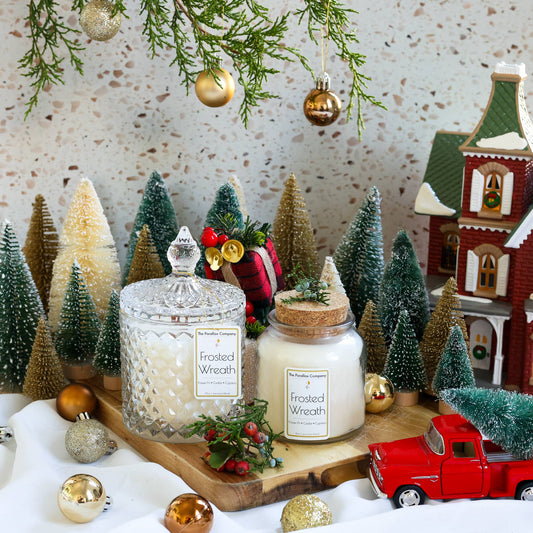 White candles labeled Frosted Wreath, surrounded by a christmas village setup, mini holiday decorations, snow-covered trees, ornaments and a red truck.